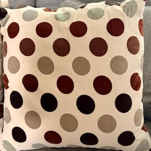 May include: A square throw pillow featuring a cream-colored background with a repeating pattern of large, circular polka dots in shades of dark brown, medium brown, light beige, and muted sage green arranged in a grid-like formation.