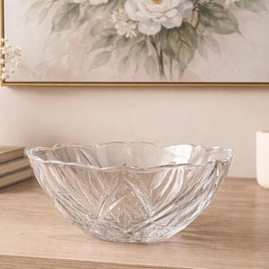 Könnte beinhalten: Eine klare Glasschale mit gewelltem Rand und Rautenmuster. Die Schale steht auf einer hellen Holzoberfläche. Im Hintergrund befindet sich ein Blumenbild.