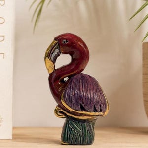 May include: A decorative flamingo figurine with a maroon head and neck, a purple body, and a green base. The beak is gold-colored, and the figurine is placed on a wooden surface.