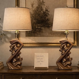 Può includere: Due lampade da tavolo abbinate con paralumi in tessuto beige. Le basi sono color bronzo con disegni floreali. Un piccolo biglietto con le parole "Good friends, Good wine, Good times" è sul tavolo.