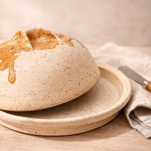May include: A ceramic butter dish with a domed lid and a matching base. The lid has a textured, speckled surface with brown accents. A small butter knife with a wooden handle rests on a linen napkin next to the dish.
