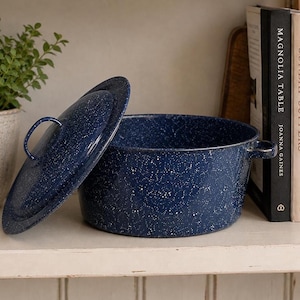 May include: A deep blue enamel pot with a white speckled pattern, featuring a matching lid with a curved handle and two side handles. The pot has a cylindrical shape and a smooth, glossy finish, presenting a classic kitchen cookware design.