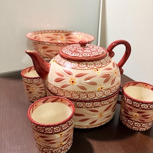 May include: A ceramic tea set with a teapot, bowl, and three cups. The set features a cream background with a floral pattern in red, brown, and yellow. The teapot has a red handle and lid. The bowl and cups have a matching design.
