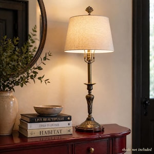 Könnte beinhalten: Eine bronzefarbene Tischlampe mit beigem Lampenschirm. Die Lampe hat einen dekorativen Fuß und einen schlanken, geschwungenen Stiel. Sie steht auf einem dunkelroten Tisch mit Büchern und einer dekorativen Schale. Die Bücher tragen die Titel "LIVE BEAUTIFULLY", "THIS IS HOME" und "HABITAT".