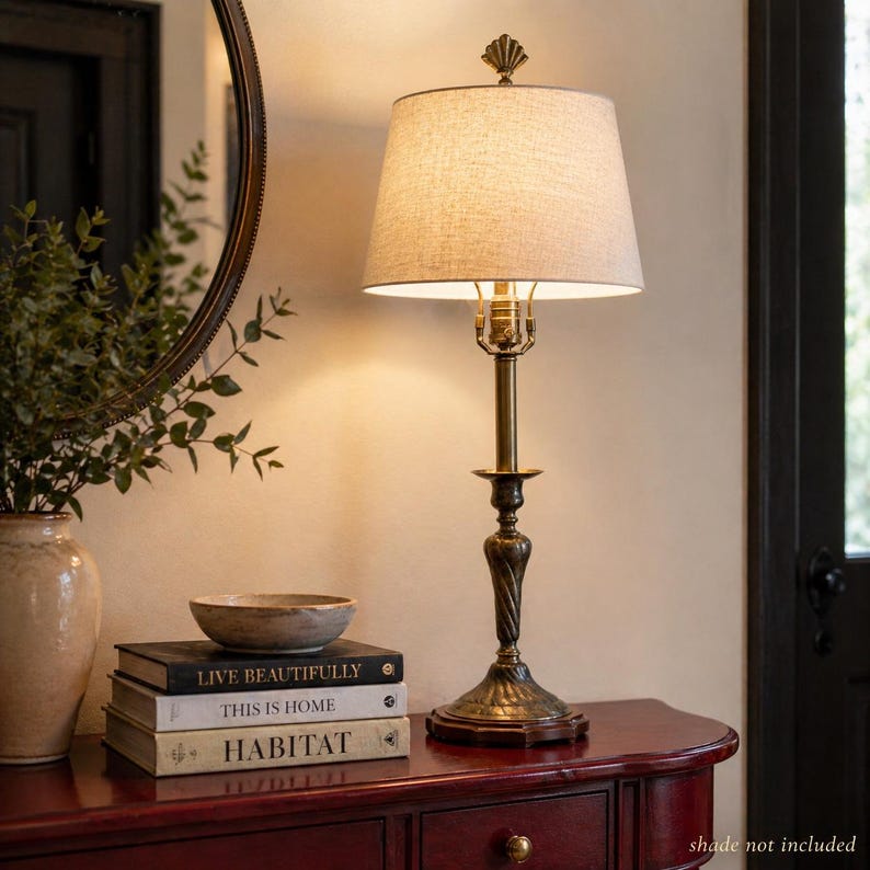 Puede incluir: Una l&aacute;mpara de mesa de color bronce con una pantalla beige. La l&aacute;mpara tiene una base decorativa y un tallo delgado y curvado. Est&aacute; sobre una mesa rojo oscuro, junto a libros y un cuenco decorativo. Los libros se titulan "LIVE BEAUTIFULLY", "THIS IS HOME" y "HABITAT".