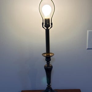 Puede incluir: Una l&aacute;mpara de mesa antigua con acabado en bronce oscuro. La l&aacute;mpara presenta una base decorativa, un tallo delgado y una bombilla encerrada en un marco de metal. La l&aacute;mpara est&aacute; sobre una superficie de madera.