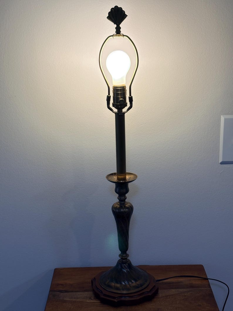 Puede incluir: Una l&aacute;mpara de mesa antigua con acabado en bronce oscuro. La l&aacute;mpara presenta una base decorativa, un tallo delgado y una bombilla encerrada en un marco de metal. La l&aacute;mpara est&aacute; sobre una superficie de madera.