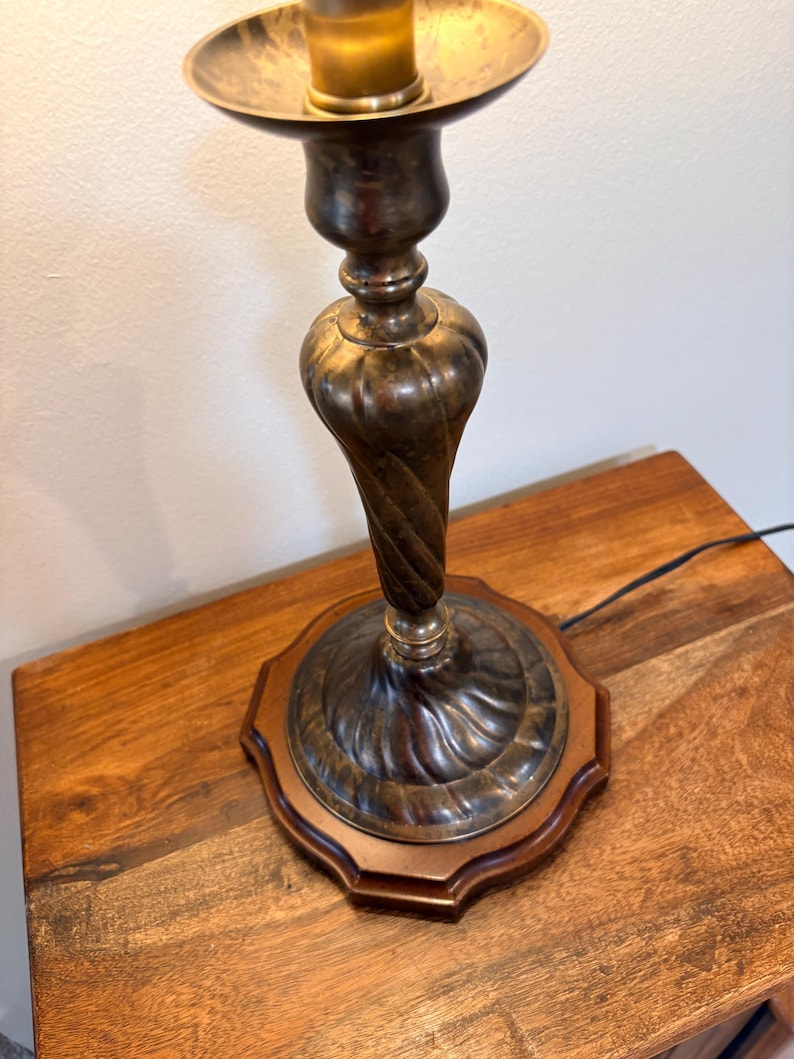 Puede incluir: Un candelero antiguo de lat&oacute;n con un dise&ntilde;o de tallo retorcido. El candelero se asienta sobre una base de madera con un borde decorativo. La parte superior del candelero tiene un plato circular para sostener una vela.
