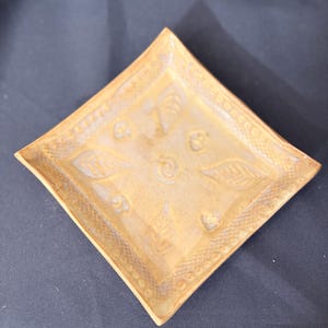 May include: A square, handmade ceramic dish with a light brown glaze. The dish features embossed leaf and swirl designs in the center and a decorative border. The corners are slightly rounded.