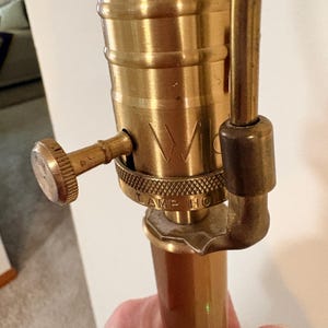 Puede incluir: Primer plano de una base de l&aacute;mpara de lat&oacute;n vintage con un acabado texturizado. La l&aacute;mpara presenta un dise&ntilde;o detallado con la palabra "W" visible. La l&aacute;mpara tiene una perilla y un cuerpo cil&iacute;ndrico, mostrando un dise&ntilde;o cl&aacute;sico.