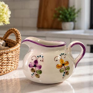 May include: A white ceramic creamer pitcher featuring a hand-painted purple rim and handle. The body is decorated with two floral motifs, one in purple and one in yellow, each with green leaves and small blue dots, set against a smooth surface.