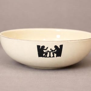 Op de afbeelding: Crèmekleurige keramische kom met een zwart silhouetontwerp van twee figuren die aan een tafel zitten. De kom heeft een afgeronde vorm en een dunne, bruine rand. Het ontwerp is gecentreerd op de buitenkant van de kom.