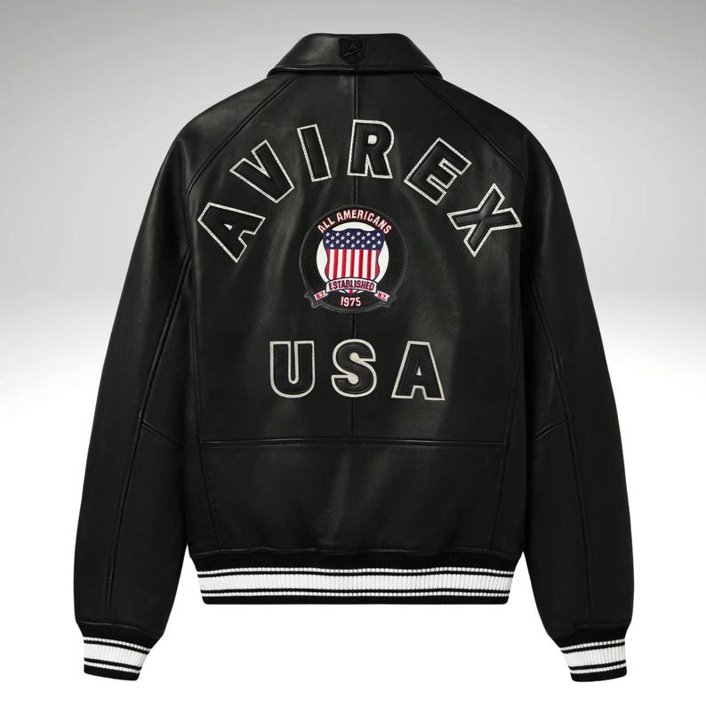 K&ouml;nnte beinhalten: Schwarze Lederjacke mit wei&szlig;en Zierleisten an den Manschetten und am Bund. Auf dem R&uuml;cken steht in wei&szlig;en Blockbuchstaben das Wort "AVIREX" und ein Aufn&auml;her mit "ALL AMERICANS ESTABLISHED 1975 USA".