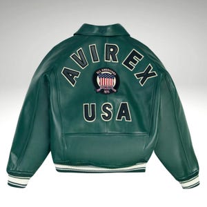 K&ouml;nnte beinhalten: Dunkelgr&uuml;ne Lederjacke mit dem Wort "AVIREX" in wei&szlig;en Buchstaben auf dem oberen R&uuml;cken. Darunter befindet sich ein Aufn&auml;her mit den Worten "All Americans" und "ESTABLISHED 1975". Das Wort "USA" ist darunter in Wei&szlig; gedruckt.