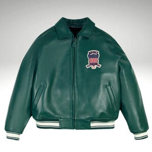 K&ouml;nnte beinhalten: Dunkelgr&uuml;ne Lederjacke im klassischen Bomberstil. Sie hat wei&szlig;-schwarz gestreifte B&uuml;ndchen und Saum. Ein gestickter Aufn&auml;her auf der Brust zeigt ein patriotisches Design mit dem Text "AVIREX".