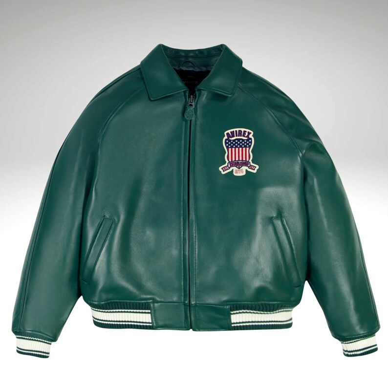 K&ouml;nnte beinhalten: Dunkelgr&uuml;ne Lederjacke im klassischen Bomberstil. Sie hat wei&szlig;-schwarz gestreifte B&uuml;ndchen und Saum. Ein gestickter Aufn&auml;her auf der Brust zeigt ein patriotisches Design mit dem Text "AVIREX".