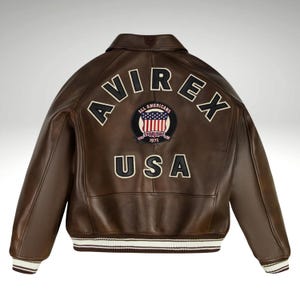 K&ouml;nnte beinhalten: Braune Lederjacke mit dem Wort "AVIREX" in wei&szlig;en Buchstaben auf dem R&uuml;cken. Ein kreisf&ouml;rmiger Aufn&auml;her mit der amerikanischen Flagge und den Worten "ALL AMERICANS ESTABLISHED 1975" ist unter dem Wort "AVIREX" zentriert. Das Wort "USA" steht darunter.