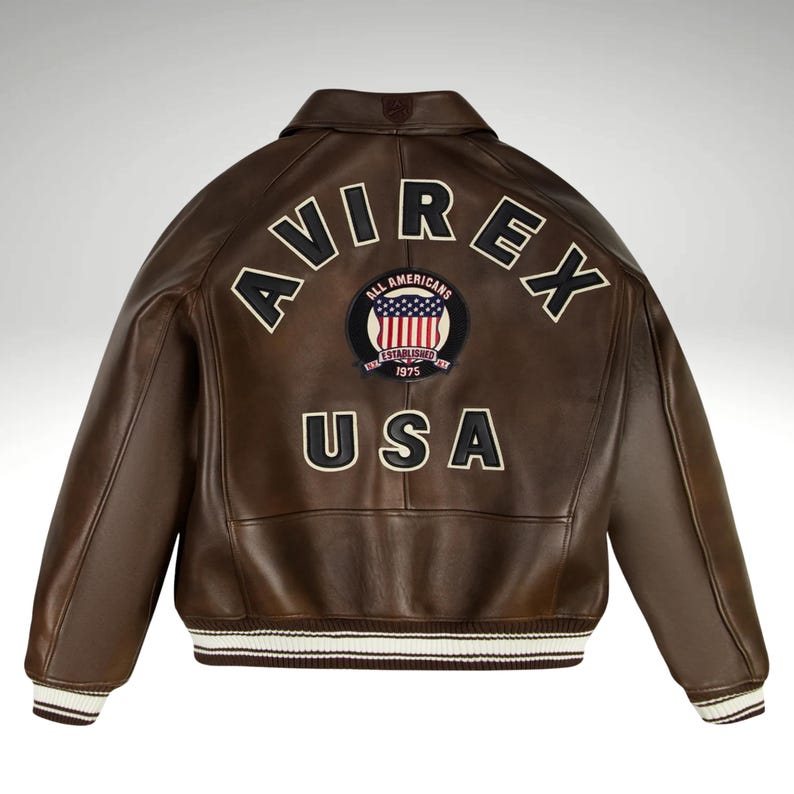 K&ouml;nnte beinhalten: Braune Lederjacke mit dem Wort "AVIREX" in wei&szlig;en Buchstaben auf dem R&uuml;cken. Ein kreisf&ouml;rmiger Aufn&auml;her mit der amerikanischen Flagge und den Worten "ALL AMERICANS ESTABLISHED 1975" ist unter dem Wort "AVIREX" zentriert. Das Wort "USA" steht darunter.