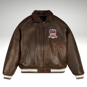 K&ouml;nnte beinhalten: Braune Lederjacke mit Rei&szlig;verschluss, Kragen und Aufn&auml;her auf der Brust. Der Aufn&auml;her zeigt das Wort "AVIREX" und ein Design in Rot, Wei&szlig; und Blau. Die Jacke hat wei&szlig;-braun gestreifte B&uuml;ndchen und Saum.