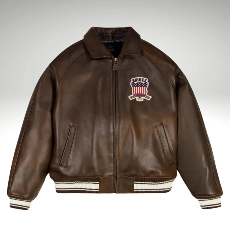 K&ouml;nnte beinhalten: Braune Lederjacke mit Rei&szlig;verschluss, Kragen und Aufn&auml;her auf der Brust. Der Aufn&auml;her zeigt das Wort "AVIREX" und ein Design in Rot, Wei&szlig; und Blau. Die Jacke hat wei&szlig;-braun gestreifte B&uuml;ndchen und Saum.