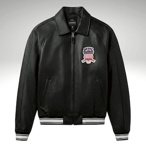 K&ouml;nnte beinhalten: Schwarze Lederjacke mit Rei&szlig;verschluss, Kragen und Aufn&auml;her auf der Brust. Die Jacke hat wei&szlig;e Streifen an den Manschetten und am Saum. Der Aufn&auml;her zeigt ein rot-wei&szlig;-blaues Design mit dem Wort "AVIREX".