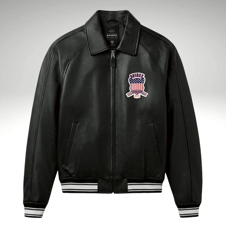 K&ouml;nnte beinhalten: Schwarze Lederjacke mit Rei&szlig;verschluss, Kragen und Aufn&auml;her auf der Brust. Die Jacke hat wei&szlig;e Streifen an den Manschetten und am Saum. Der Aufn&auml;her zeigt ein rot-wei&szlig;-blaues Design mit dem Wort "AVIREX".