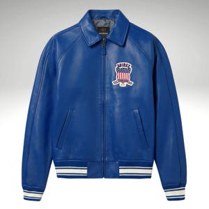 K&ouml;nnte beinhalten: Eine leuchtend blaue Lederjacke mit klassischem Kragen und Rei&szlig;verschluss. Sie hat einen Aufn&auml;her mit einem amerikanischen Flaggen-Design und dem Markennamen. Die Jacke hat wei&szlig;-blaue gestreifte B&uuml;ndchen und Saum.