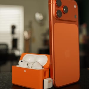 Könnte beinhalten: Orangefarbenes Smartphone und kabellose Ohrhörer mit passendem Ladecase. Das Telefon hat drei Kameralinsen und ein weißes Apple-Logo. Die Ohrhörer sind weiß und sitzen in einem offenen orangefarbenen Case. Das Case hat eine weiße "del"-Taste.