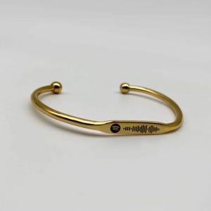 May include: A gold-colored cuff bracelet with a Spotify code engraved on the front. The bracelet has rounded ball ends and a smooth, polished finish. The Spotify code is black and is a series of vertical lines.