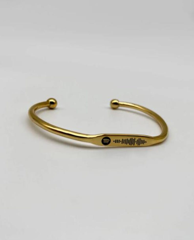 May include: A gold-colored cuff bracelet with a Spotify code engraved on the front. The bracelet has rounded ball ends and a smooth, polished finish. The Spotify code is black and is a series of vertical lines.