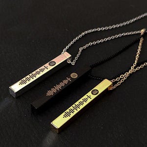 Puede incluir: Tres collares con colgantes en forma de barra en oro, plata y negro. Cada colgante presenta un patrón de onda de sonido único y un logotipo circular. Los collares cuelgan de delicadas cadenas, sobre un fondo oscuro.
