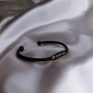May include: Black cuff bracelet with a gold Spotify code and logo. The bracelet is made of a curved, solid band with rounded ends. The Spotify code is a series of vertical lines. The bracelet is displayed on a silky, gray surface.