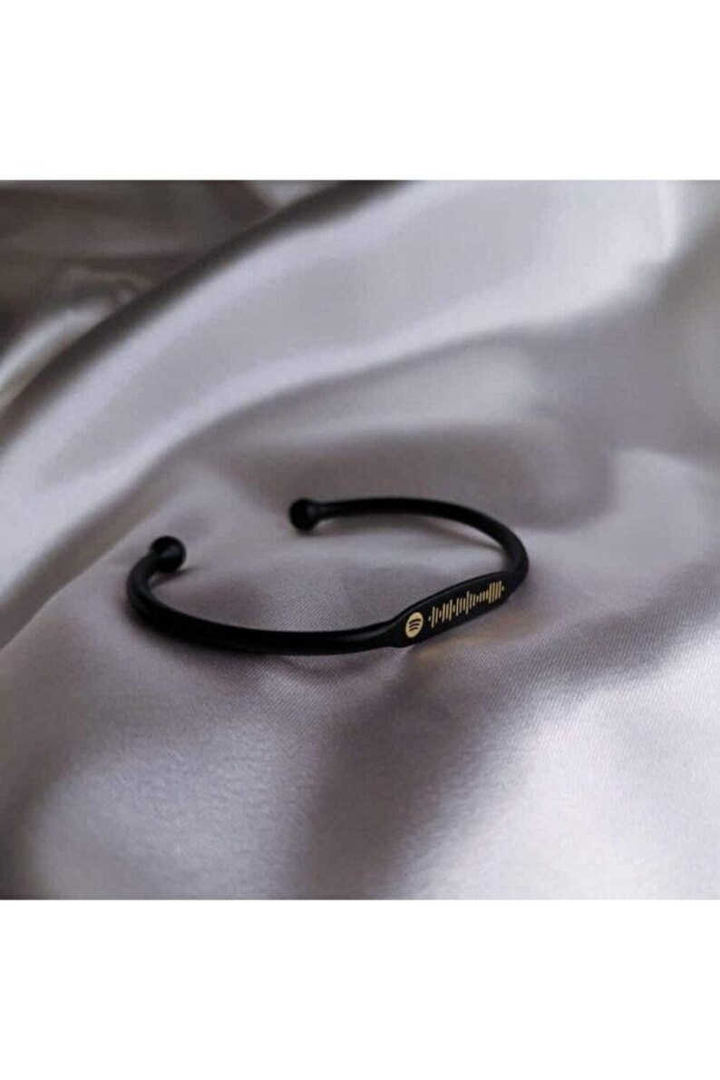 May include: Black cuff bracelet with a gold Spotify code and logo. The bracelet is made of a curved, solid band with rounded ends. The Spotify code is a series of vertical lines. The bracelet is displayed on a silky, gray surface.