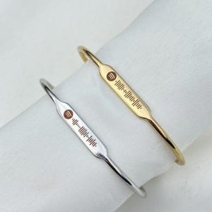 May include: Two metal Spotify code bracelets. One is gold-toned, the other is silver-toned. Each bracelet has a rectangular plate with a Spotify logo and a series of lines representing a song's code. The bracelets are displayed on a white surface.