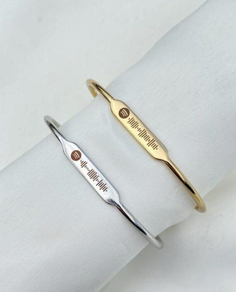 May include: Two metal Spotify code bracelets. One is gold-toned, the other is silver-toned. Each bracelet has a rectangular plate with a Spotify logo and a series of lines representing a song's code. The bracelets are displayed on a white surface.