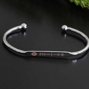 May include: A silver-colored cuff bracelet with a Spotify code engraved on a rectangular plate. The bracelet has rounded ball ends and is displayed on a dark surface. The Spotify code is in a light brown color.