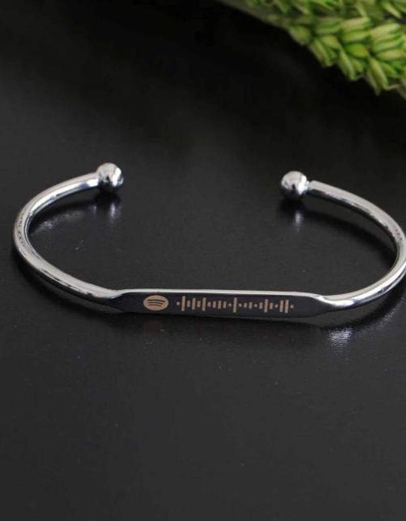 May include: A silver-colored cuff bracelet with a Spotify code engraved on a rectangular plate. The bracelet has rounded ball ends and is displayed on a dark surface. The Spotify code is in a light brown color.