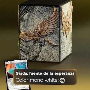 Puede incluir: Una caja de almacenamiento de cartas negra y gris con un relieve detallado de un ángel con alas extendidas. La caja tiene el texto "Giada, fuente de la esperanza" y "Color mono white:" con un símbolo de maná blanco. Se ve una carta.