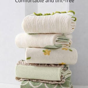 Peut inclure: Une pile de couvertures pliées, dans des tons crème et verts. Certaines couvertures présentent des motifs floraux ou des bordures décoratives. Le texte "Durability & Anti-Pilling" et "Comfortable and lint-free" est visible.