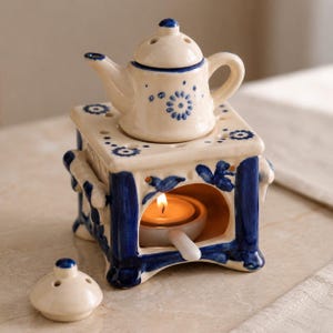 May include: A ceramic teapot-shaped oil warmer with a blue and white floral design. The warmer has a small teapot on top and a candle holder inside. The design includes blue floral patterns and accents. The lid is detached and placed in front.