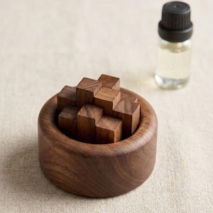 May include: A wooden puzzle with small, square blocks inside a round, wooden bowl. A small glass bottle with a black cap is next to the puzzle. The items are on a beige surface.