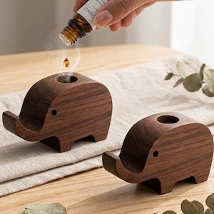 May include: Two wooden elephant-shaped essential oil diffusers. The diffusers are dark brown with a smooth finish and a small hole on top. A hand is pouring oil from a bottle labeled "essence" into one of the diffusers. The diffusers are on a wooden table.