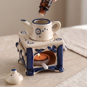 May include: A ceramic essential oil diffuser with a teapot design, featuring blue floral patterns and a small candle inside. A hand is pouring oil from a brown bottle labeled "DMA AROMANCE OIL". The diffuser is on a patterned surface.