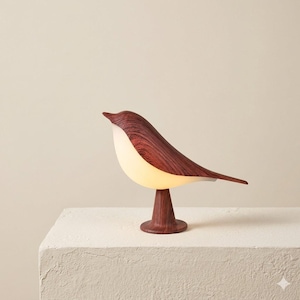 May include: A decorative bird-shaped lamp with a warm, inviting glow. The lamp features a wooden base and body with a reddish-brown finish, and a white, illuminated belly. The design is modern and minimalist, suitable for home decor.