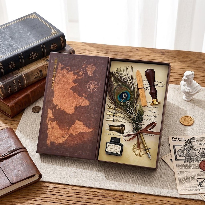 Op de afbeelding: Een vintage schrijfgarnituur in een decoratieve doos. De set bevat een ganzenveer met een pauwenveer, een inktfles, een zegel en een klein notitieboekje. De doos heeft een kaartontwerp op de omslag en is omgeven door antieke boeken.