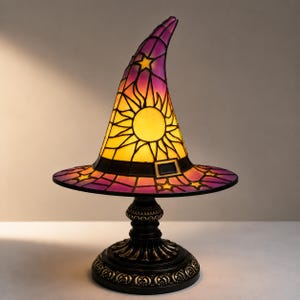 May include: A stained glass table lamp shaped like a pointed witch hat. The lamp features a yellow sun design on the cone, surrounded by purple glass with star cutouts. It has a black band with a buckle and sits on a dark, ornate metal base.