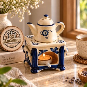 May include: A cream-colored ceramic oil warmer featuring a blue floral pattern and a matching teapot-shaped top. The square base has an arched opening containing a small white tea light candle holder with a lit candle inside the ceramic structure.