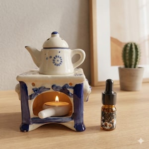 May include: A ceramic teapot-shaped essential oil warmer with blue floral accents sits atop a square base. A lit candle is inside the warmer. A small glass bottle with a black dropper is next to the warmer.