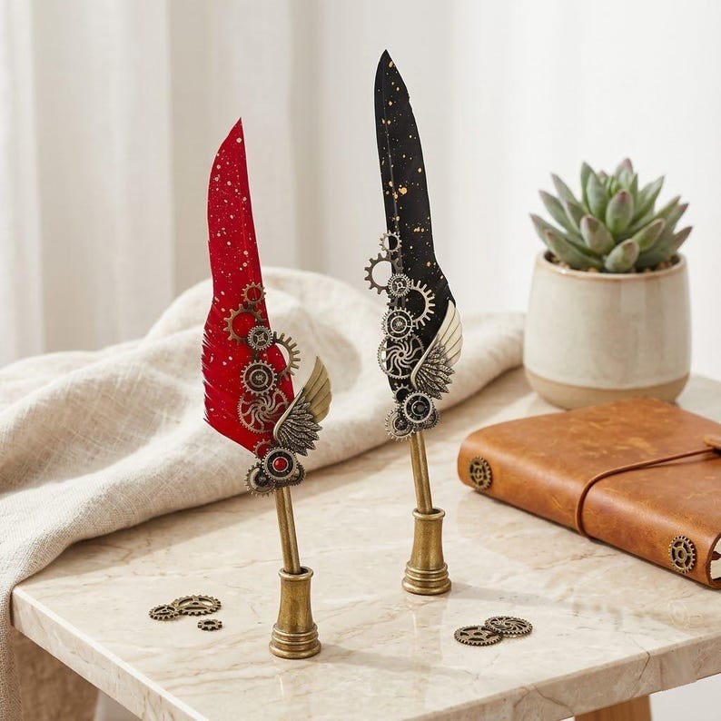 Op de afbeelding: Twee decoratieve vulpennen in steampunk-stijl. De ene is rood met gouden accenten, de andere zwart met gouden spikkels. Beide pennen hebben tandwiel- en vleugelversieringen en zitten in messingkleurige penhouders.