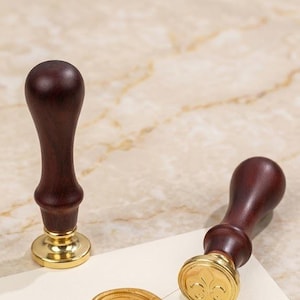 Op de afbeelding: Twee houten lakstempels met goudkleurige bases en donkerbruine handvatten. E&eacute;n stempel staat rechtop, de andere ligt op een cr&egrave;mekleurige envelop met een gouden lakzegel en roze lint. De zegel heeft een bloemenmotief.